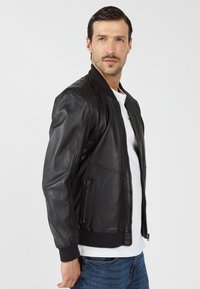 Zwarte leren bomberjack met een geribbelde kraag, rits aan de voorkant, zijzakken en gestructureerde mouwen, gedragen over een witte t-shirt.