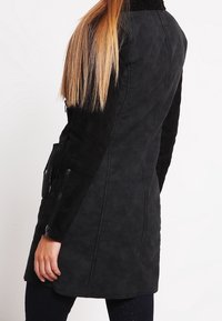 Cappotto in faux suede nero con finitura testurizzata, dotato di maniche lunghe, tasche laterali e chiusura con zip. Design liscio e aderente.