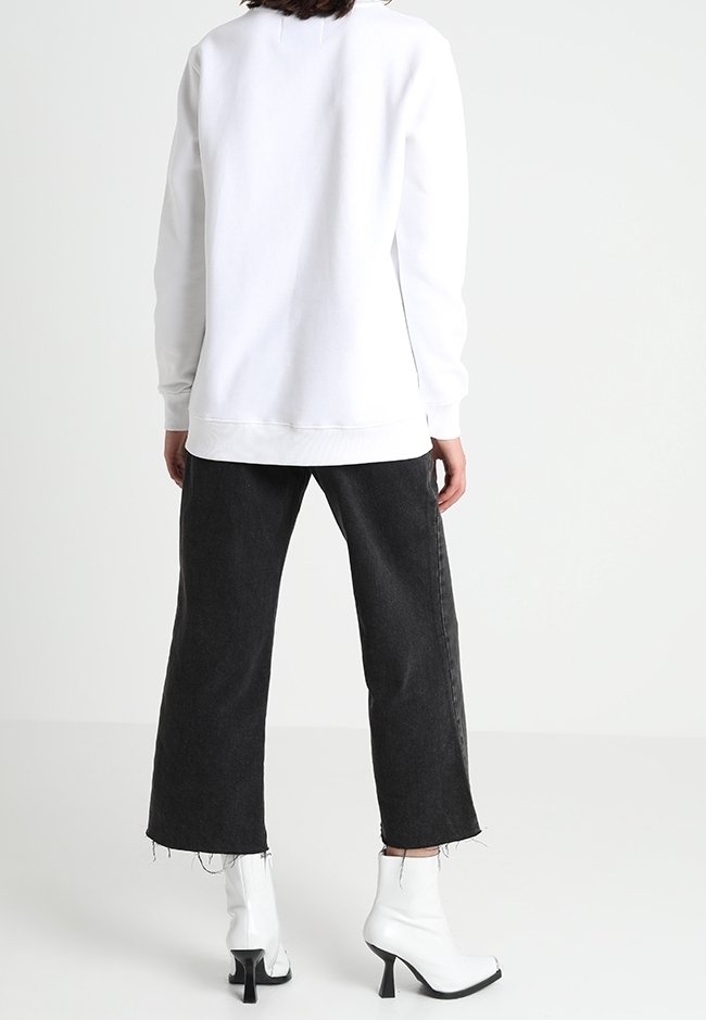 Personne portant un haut blanc à manches longues, un jean noir large raccourci avec des ourlets effilochés, et des bottines blanches à talons carrés, debout de dos.