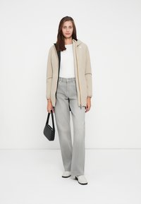Beige ritsjack met zwarte voering, gecombineerd met een witte blouse en grijze wijde broek. Accessoires zijn een kleine zwarte tas en witte schoenen.