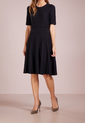 Femme portant une robe noire plissée longueur genou avec manches courtes et des chaussures à talons taupe, debout devant un fond uni.