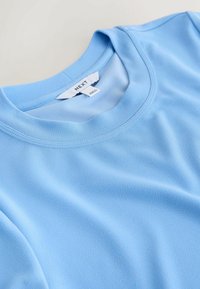 Haut bleu clair col ras du cou en tissu finement texturé, soigneusement plié, montrant l'étiquette de la marque "NEXT" cousue à l'intérieur du col.