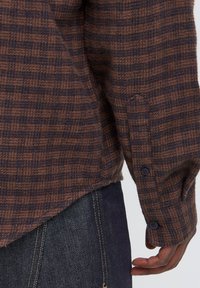 Poignet de chemise à manches longues à carreaux marron et noir avec boutons noirs portée sur un jean bleu foncé, montrant la main détendue d'une personne le long du corps.