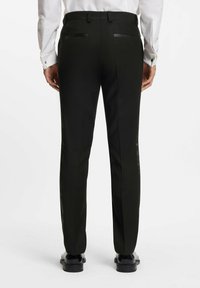 Uomo in pantaloni neri, camicia bianca con maniche arrotolate e scarpe eleganti nere lucide, in piedi di fronte a uno sfondo bianco semplice.