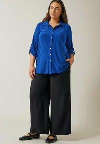 Camicia blu a maniche tre quarti con due tasche sul petto, abbinata a pantaloni neri a gamba larga e ballerine nere con borchie.