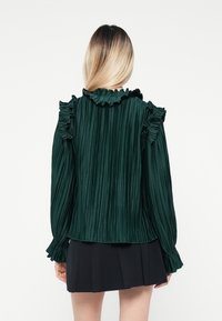 Blusa plissettata verde scuro con scollatura e polsini arricciati, caratterizzata da maniche lunghe e una finitura testurizzata, abbinata a una gonna nera.