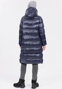 Marinblå uppblåsbar jacka med huva, som har en lång, cylindrisk design och horisontellt quiltat mönster. Bärs över grå leggings och svarta stövlar.