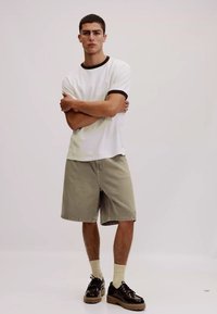 T-shirt blanc à manches courtes avec col noir ; short vert clair ; chaussures noires avec semelle épaisse beige ; chaussettes beige de hauteur moyenne.