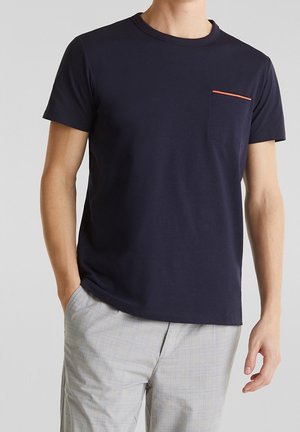 Uomo che indossa una t-shirt blu navy a maniche corte con una piccola tasca sul petto bordata di arancione e pantaloni grigio chiaro a quadri.