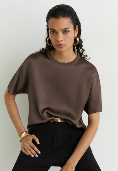 Woman with dark curly hair wearing a brown short-sleeve top, black pants, gold hoop earrings, and gold bracelets stands against a white background.
