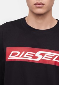 Νέος άνθρωπος που φοράει μαύρο T-shirt με το κόκκινο και λευκό λογότυπο Diesel στο στήθος, εμφανίζοντας μόνο το κάτω μέρος του προσώπου και τον λαιμό.
