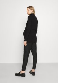 Svart strikket topp med høy hals og snøring, kombinert med svarte striperbukser. Modellen har på seg svarte loafers; minimal tilbehør.