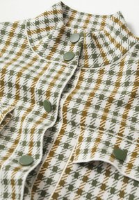 Tkanina we wzór houndstooth w odcieniach zieleni i brązu, z wysokim kołnierzem, ozdobiona okrągłymi zielonymi guzikami oraz teksturowanymi detalami plecionki.