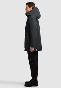 Czarna, ocieplana parka z wysokim kołnierzem, dużym kapturem, kieszeniami bocznymi i czarnymi spodniami cargo. Teksturowane czarne buty dopełniają całość.