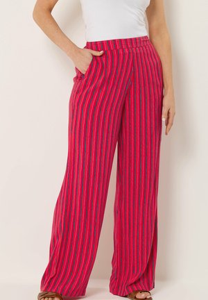 Donna che indossa pantaloni rossi a vita alta e gamba larga con strisce verticali blu e rosa, abbinati a una maglietta bianca e sandali marroni.