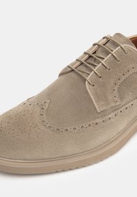 Chaussure habillée en daim beige avec des détails perforés, des lacets fins et une semelle en caoutchouc léger ; bout rond et texture lisse.