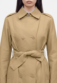 Femme portant un trench-coat beige à double boutonnage avec une ceinture à la taille nouée en nœud et une chemise bleu clair à col en dessous.
