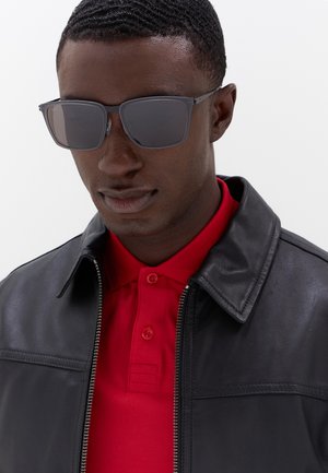 Uomo con occhiali da sole scuri, giacca di pelle nera e camicia rossa con colletto su uno sfondo bianco semplice.