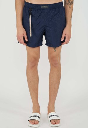 Homme portant un short de bain bleu marine avec une étiquette latérale et des sandales blanches Emporio Armani, montrant des tatouages sur les bras et la jambe.