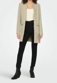 Beżowy długi żakiet z kołnierzem, przednimi suwakami i bocznymi kieszeniami, w zestawieniu z czarnymi dopasowanymi spodniami i botkami za kostkę. Gładka faktura materiału.