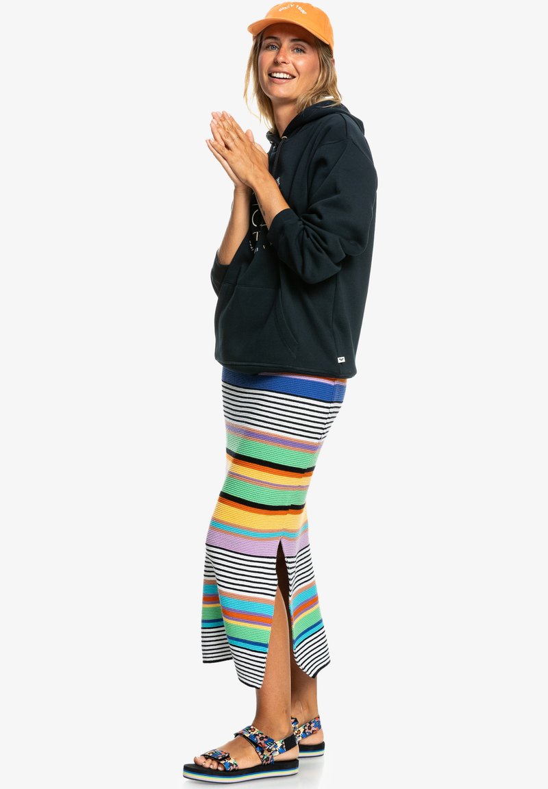 Black hoodie, multicolored striped knit skirt with side slit, and patterned sandals. Bright orange cap. Casual and layered outfit.