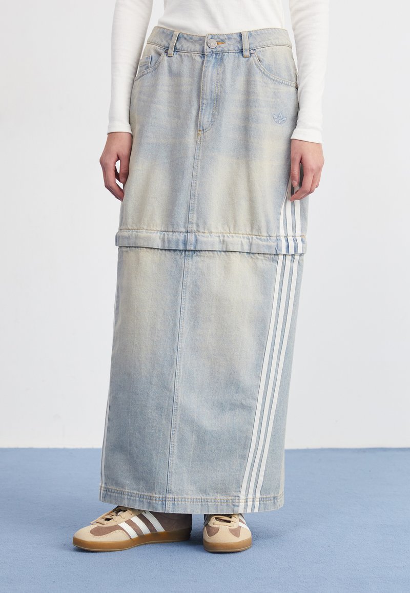 Jupe maxi en denim bleu clair avec une finition délavée, ornée de deux rayures verticales blanches et de poches à l'avant. Associée à des baskets marron et beige.