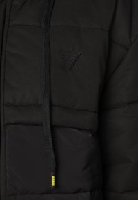 Giacca a vento nera con texture trapuntata, dotata di cappuccio, coulisse e una patch con logo triangolare sul petto. Tasca frontale visibile.