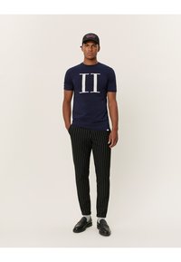Camiseta azul marino con un gran diseño blanco de "II", combinada con pantalones negros de rayas verticales y zapatos negros. El modelo lleva una gorra negra con texto rosa.