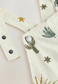 Close-up van crèmekleurige stofoveralls met groene cactus- en sterrenprints, verstelbare riemen met zwarte drukknopen en metalen nitdetail.