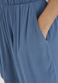 Niebieskie szorty z teksturą, plisami i bocznymi kieszeniami, wykonane z miękkiego, lekkiego materiału. Zdjęcie zbliżeniowe podkreśla detale kieszeni.