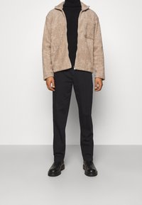 Beige, fuzzy jakke med lynlås foran og høj krave, parret med sorte bukser og sorte ankelstøvler. Har detaljer med frontlomme.