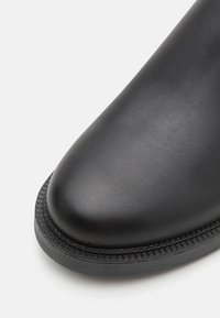 Boot negru din piele cu vârful rotund, suprafață texturată și talpă groasă din cauciuc, având un detaliu cusut pe margine.