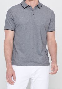 Hombre vistiendo una camiseta polo gris de manga corta con cuello y bordes de mangas a rayas negras, combinado con pantalones blancos, de pie contra un fondo liso.