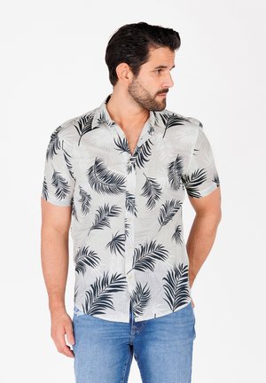 Korte mouwen overhemd van lichte stof, met een witte achtergrond en zwarte palmbladpatronen. Gecombineerd met blauwe jeans.