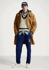 Ung man i en beige duffle-kappa, lager av stickade tröjor och rutiga skjortor, marinblå sweatpants med USA-logotyp, vita sneakers och en blå "Ralph" baseballkeps.