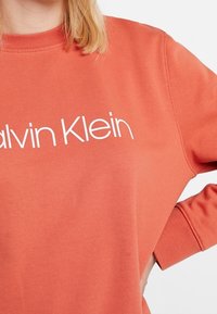 Felpa arancione con scollo rotondo, con logo "Calvin Klein" bianco stampato sul petto. Materiale morbido e vestibilità comoda.