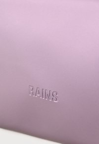 Λείο ματ μοβ επιφάνεια με τη λέξη "RAINS" ανάγλυφη σε κεφαλαία γράμματα κοντά στο κέντρο στο κάτω μέρος.