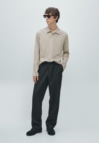 Beige poloshirt met lange mouwen en kraag, gecombineerd met een donkere broek en zwarte schoenen. De stof oogt zacht met een gladde textuur.