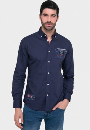 Camisa azul marino de botones hecha de algodón con un diseño ajustado. Presenta logotipos bordados y acentos en rojo y blanco, y puños abotonados.