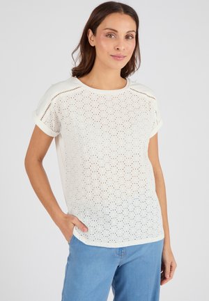 Haut blanc à manches courtes en tissu perforé, col rond, avec des poignets roulés. Associé à un jean bleu clair. Texture lisse, coupe décontractée.