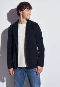 Black corduroy blazer with a tailored fit, notched lapels, and two front pockets, worn over a light-colored t-shirt and denim jeans.