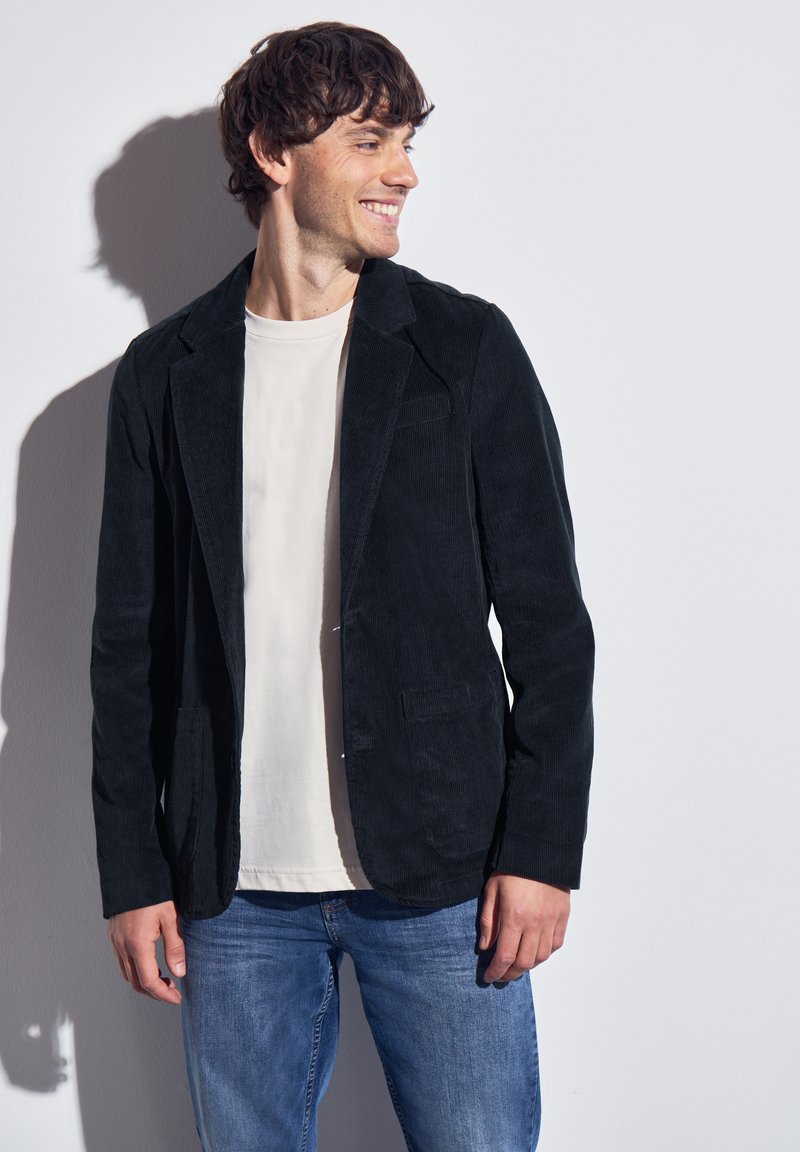 Black corduroy blazer with a tailored fit, notched lapels, and two front pockets, worn over a light-colored t-shirt and denim jeans.