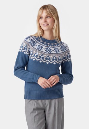 Donna che indossa un maglione blu lavorato a maglia con motivi di fiocchi di neve bianchi e grigi e motivi geometrici, abbinato a pantaloni grigio chiaro, in piedi con le mani giunte.