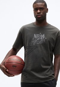 Νέος άνδρας που κρατάει μια μπάλα μπάσκετ Spalding, φορώντας ένα σκουρόχρωμο γκρι μπλουζάκι NBA All-Star, μπροστά σε ένα απλό λευκό φόντο.