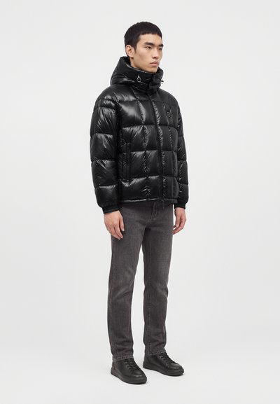 Jeune homme portant une doudoune noire brillante avec capuche, un jean gris et des baskets noires, debout devant un fond clair uni.