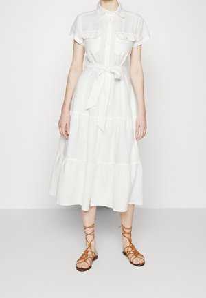 Kobieta ubrana w białą, midi sukienkę na krótki rękaw z guzikami i przewiązanym w pasie paskiem oraz brązowe, wiązane sandały na płaskim obcasie, stojąca na jednolitym tle.