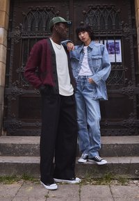 Two individuals stand on stone steps. One wears a burgundy and gray cardigan, white shirt, and black pants. The other is in a light blue denim jacket and jeans, both in black sneakers.