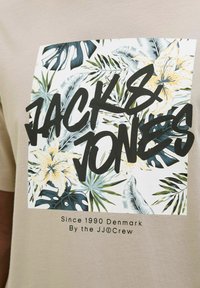 Camiseta beige con un gráfico de hojas tropicales y flores amarillas, texto negro en negrita que dice "JACK & JONES" y texto más pequeño debajo.