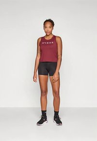 Μπορντό αμάνικο αθλητικό τοπ με το λογότυπο "HYROX", συνδυασμένο με μαύρο σορτς και μαύρα αθλητικά παπούτσια. Μαλλιά δεμένα σε αλογοουρά, στέκεται με τα χέρια χαλαρά.