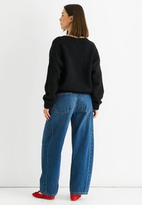 Černý pletený svetr se spadlými rameny, doplněný o světle modré volné džíny s zadními kapsami. Červené boty dokončují tento outfit.